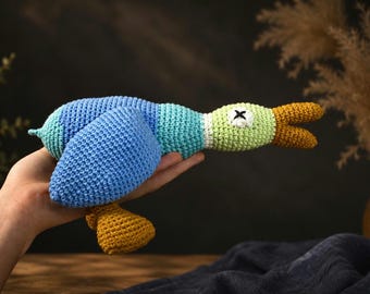 May include: A crocheted duck toy featuring a blue body, a light green neck, and a white ring around the neck. The duck has a yellow beak and yellow feet, with a white eye patch featuring a black stitched X to represent the eye. The texture is yarn.