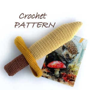 May include: A crocheted sword toy with a brown handle and a yellow blade. The sword is lying on a white surface with a picture of a hedgehog and a mushroom.