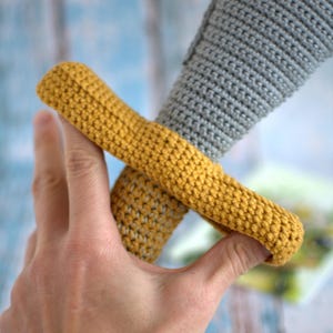 May include: A crocheted toy sword with a gray handle and a yellow guard. The sword is held in a hand.