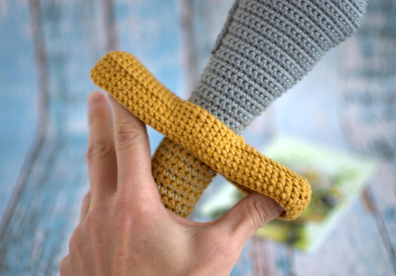 May include: A crocheted toy sword with a gray handle and a yellow guard. The sword is held in a hand.
