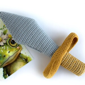 May include: A crocheted toy sword with a gray blade and a yellow handle. The sword is lying on a white surface next to a card with a cartoon frog wearing a flower crown.