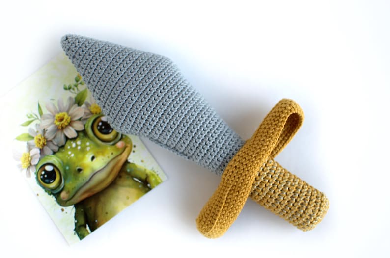 May include: A crocheted toy sword with a gray blade and a yellow handle. The sword is lying on a white surface next to a card with a cartoon frog wearing a flower crown.