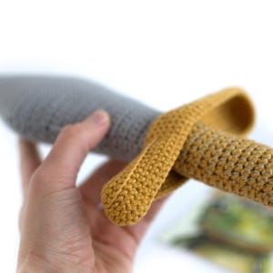 May include: A crocheted toy sword with a gray blade and a yellow handle. The sword is held in a hand.