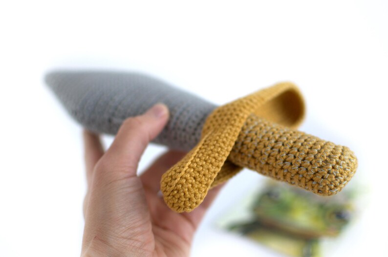 May include: A crocheted toy sword with a gray blade and a yellow handle. The sword is held in a hand.