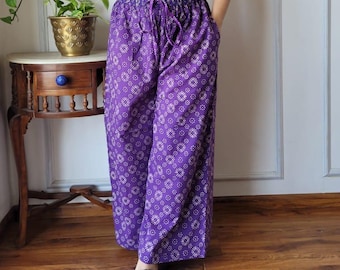 May include: A woman wearing a white crop top and purple palazzo pants with a floral pattern. The pants have a drawstring waist. The outfit is paired with green sandals. The background includes a wooden floor and a white wall.