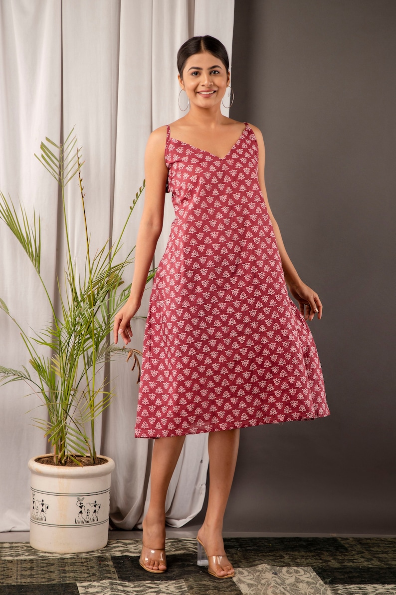 K&ouml;nnte beinhalten: Eine Frau tr&auml;gt ein maronfarbenes, &auml;rmelloses Midikleid mit wei&szlig;em Blumenmuster. Das Kleid hat einen V-Ausschnitt und eine lockere Passform. Sie tr&auml;gt transparente Sandalen mit Absatz.