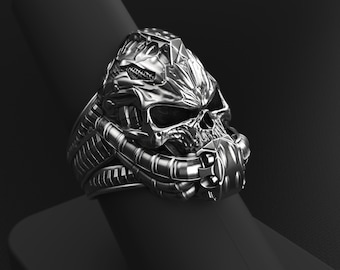May include: A silver-colored ring with a skull design. The skull has intricate details and is surrounded by mechanical-looking elements. The ring is set against a dark background, highlighting its metallic sheen.