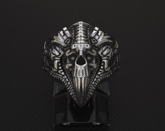 May include: A silver ring with a detailed skull design. The skull has a pointed nose and two horns on the top. The ring is set on a black background.