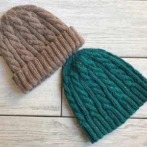 Op de afbeelding: Twee gebreide mutsen, een bruine en een groene, met een kabelbreipatroon. De mutsen liggen op een wit houten oppervlak.