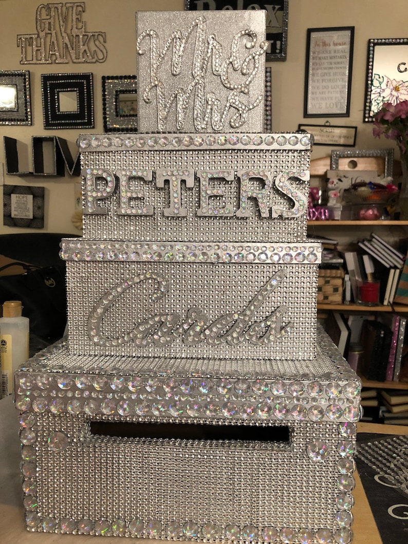 Puede incluir: Una caja de tarjetas de boda plateada con un dise&ntilde;o en capas. La caja est&aacute; cubierta de pedrer&iacute;a y presenta los nombres "Mrs & Mrs", "Peters" y "Candice". La caja tiene una ranura para tarjetas y est&aacute; decorada con detalles brillantes.