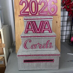 Puede incluir: Una caja de tarjetas de tres niveles con purpurina plateada y detalles en rosa. La parte superior de la caja tiene el a&ntilde;o "2026" en rosa, y el nivel medio tiene el nombre "AVA". El nivel inferior dice "Tarjetas".