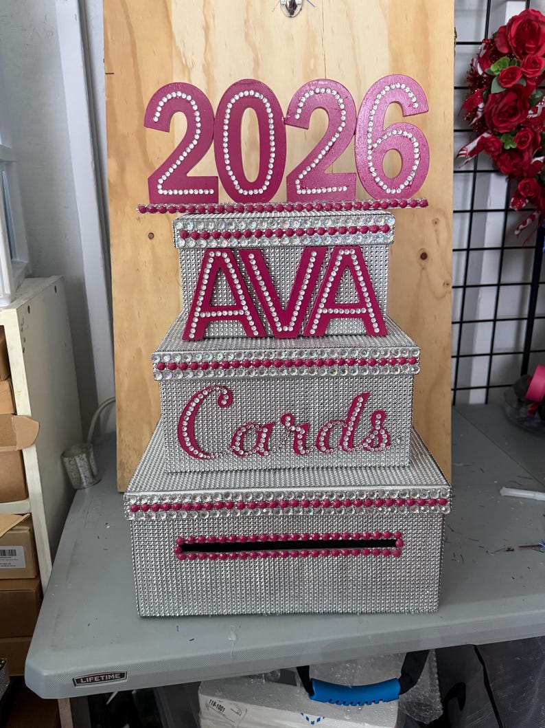 Puede incluir: Una caja de tarjetas de tres niveles con purpurina plateada y detalles en rosa. La parte superior de la caja tiene el a&ntilde;o "2026" en rosa, y el nivel medio tiene el nombre "AVA". El nivel inferior dice "Tarjetas".