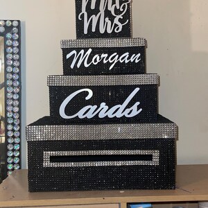 Puede incluir: Una caja de tarjetas negra de varios niveles con ribete de diamantes de imitaci&oacute;n plateados. La caja est&aacute; decorada con las palabras "Mrs & Mrs", "Morgan" y "Cards" en escritura blanca. La caja est&aacute; dise&ntilde;ada para parecerse a una tarta de boda.