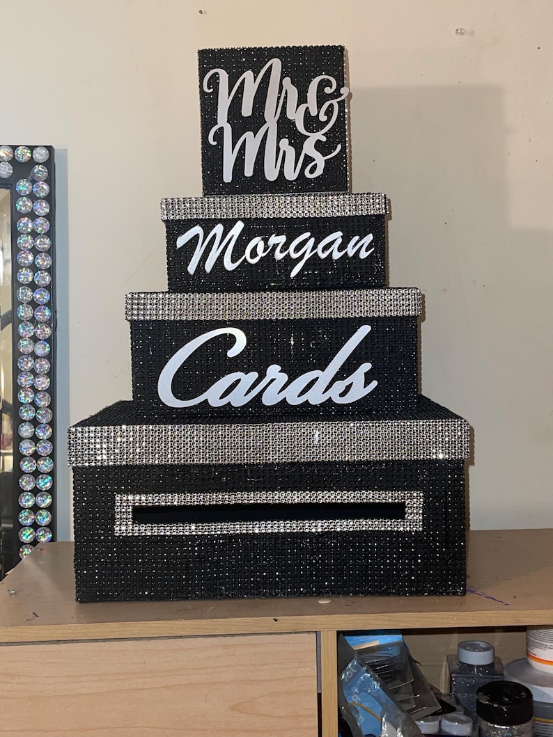 Puede incluir: Una caja de tarjetas negra de varios niveles con ribete de diamantes de imitaci&oacute;n plateados. La caja est&aacute; decorada con las palabras "Mrs & Mrs", "Morgan" y "Cards" en escritura blanca. La caja est&aacute; dise&ntilde;ada para parecerse a una tarta de boda.
