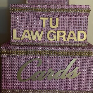 Puede incluir: Una caja de tarjetas de varios niveles, de color lavanda, incrustada con pedrer&iacute;a. La caja presenta letras doradas que dicen "TU LAW GRAD" y "Cards". Los bordes dorados acent&uacute;an los bordes, creando un dise&ntilde;o elegante y festivo.