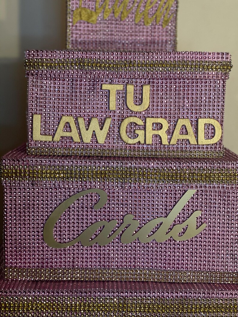 Puede incluir: Una caja de tarjetas de varios niveles, de color lavanda, incrustada con pedrer&iacute;a. La caja presenta letras doradas que dicen "TU LAW GRAD" y "Cards". Los bordes dorados acent&uacute;an los bordes, creando un dise&ntilde;o elegante y festivo.