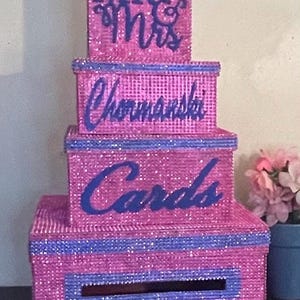 Puede incluir: Una caja de tarjetas de varios niveles, cubierta de purpurina rosa con detalles en azul. La parte superior dice "MR & Mrs", el segundo nivel tiene el nombre "Chormanski" y el tercer nivel dice "Cards". Una ranura es visible en la parte delantera del nivel inferior.