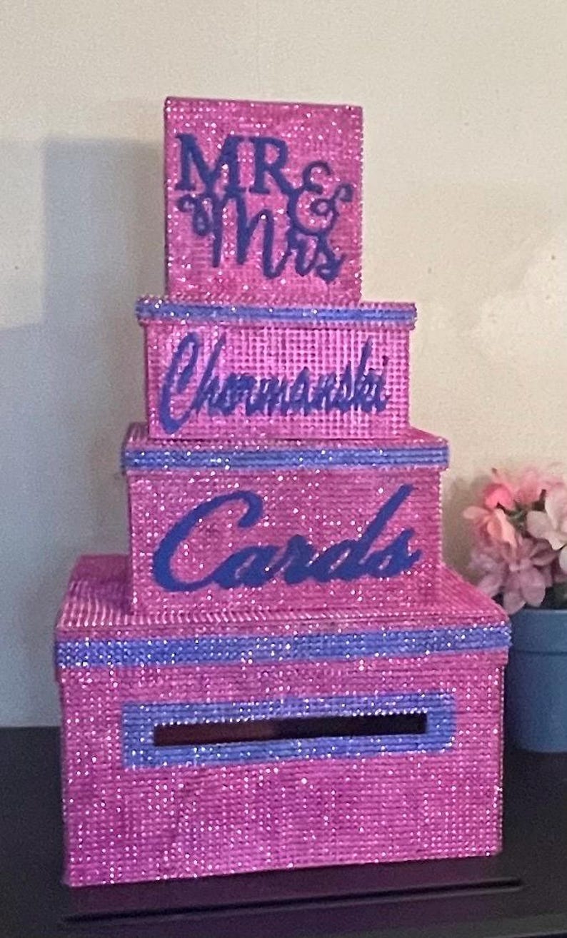 Puede incluir: Una caja de tarjetas de varios niveles, cubierta de purpurina rosa con detalles en azul. La parte superior dice "MR & Mrs", el segundo nivel tiene el nombre "Chormanski" y el tercer nivel dice "Cards". Una ranura es visible en la parte delantera del nivel inferior.