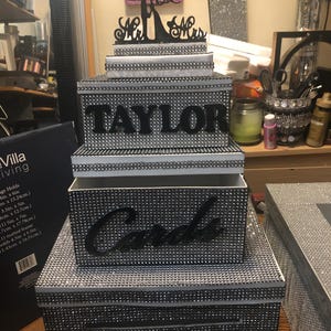 Puede incluir: Una caja de tarjetas de boda de tres niveles con un exterior cubierto de pedrer&iacute;a. El nivel superior tiene un adorno negro de Sr. y Sra. El nivel medio tiene el nombre "TAYLOR" en letras negras. El nivel inferior dice "Cards".