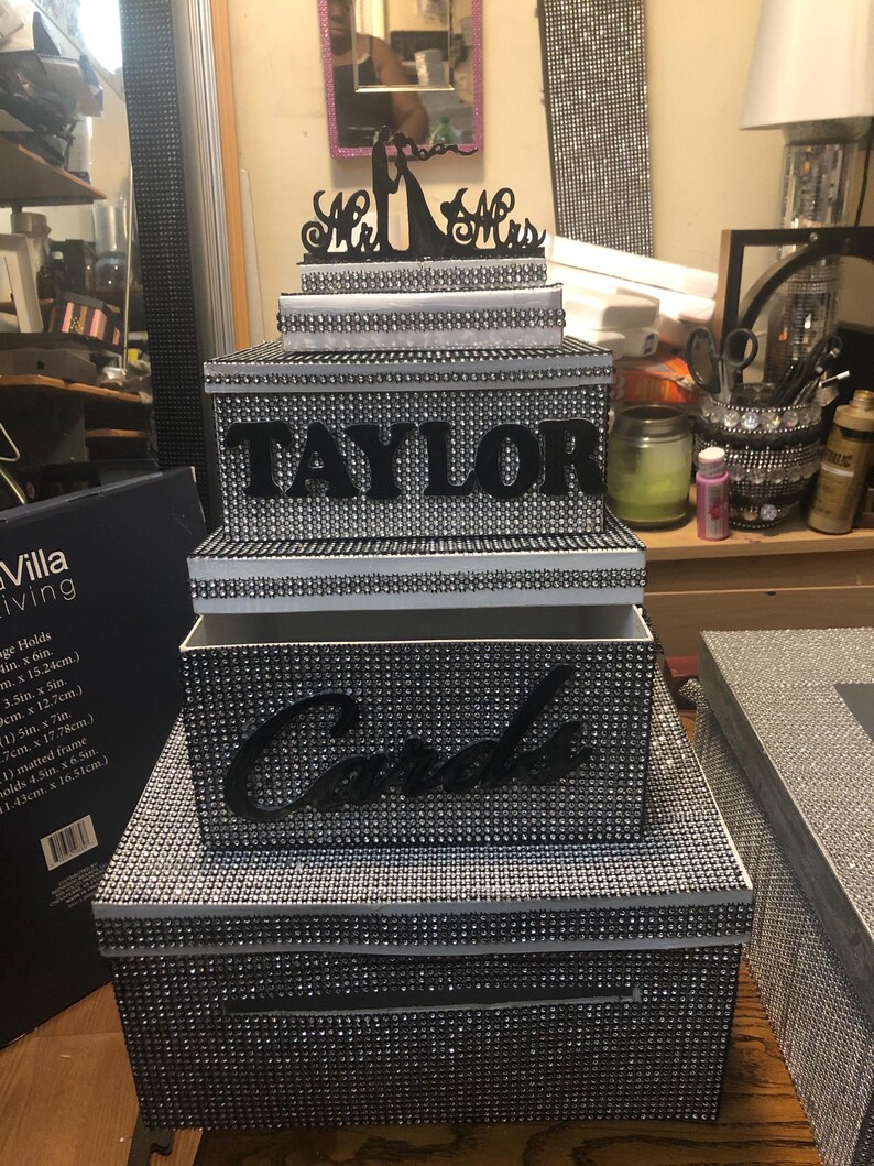 Puede incluir: Una caja de tarjetas de boda de tres niveles con un exterior cubierto de pedrer&iacute;a. El nivel superior tiene un adorno negro de Sr. y Sra. El nivel medio tiene el nombre "TAYLOR" en letras negras. El nivel inferior dice "Cards".