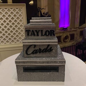 Puede incluir: Una caja de tarjetas de varios niveles, cubierta de pedrer&iacute;a plateada, con el nombre "TAYLOR" en letras negras. La caja tiene una ranura para tarjetas y est&aacute; coronada con una silueta negra de una novia y un novio con "Mr & Mrs".