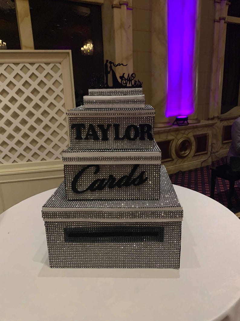 Puede incluir: Una caja de tarjetas de varios niveles, cubierta de pedrer&iacute;a plateada, con el nombre "TAYLOR" en letras negras. La caja tiene una ranura para tarjetas y est&aacute; coronada con una silueta negra de una novia y un novio con "Mr & Mrs".