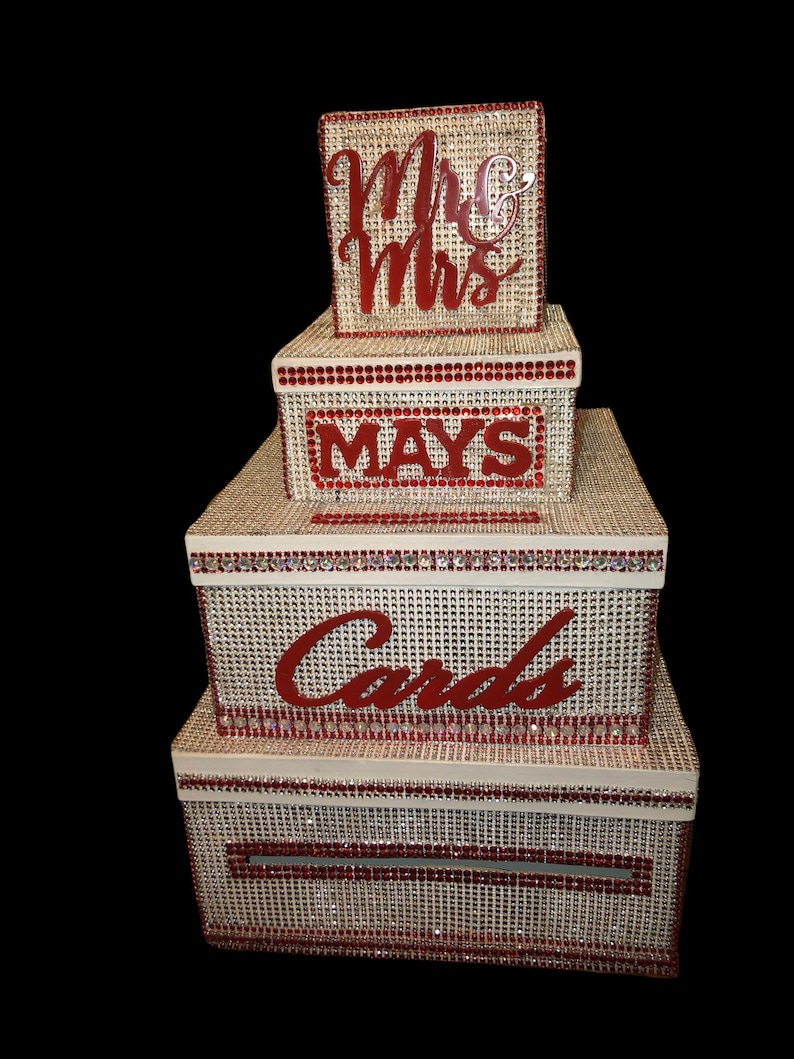 Puede incluir: Un juego de cajas de tarjetas de boda apiladas con una textura tejida beige, adornadas con pedrer&iacute;a roja. La caja superior dice "Mr & Mrs", la caja del medio "MAYS" y la caja m&aacute;s grande "Cards".