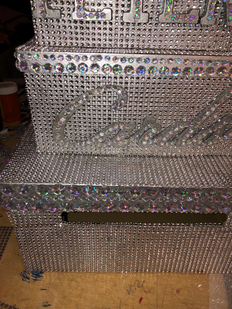 Puede incluir: Una caja plateada y adornada con pedrer&iacute;a con la palabra "Cash" escrita con letras claras y brillantes. La caja est&aacute; cubierta de peque&ntilde;os y brillantes pedrer&iacute;a y tiene una ranura en la parte delantera. Los bordes est&aacute;n forrados con gemas iridiscentes m&aacute;s grandes.