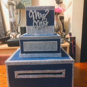 Puede incluir: Una caja de tarjetas de boda azul marino de tres niveles con ribete de diamantes de imitaci&oacute;n plateados. El nivel superior tiene "Mrs & Mrs" en escritura plateada. El nivel medio tiene el nombre "HOFFMAN" en plata.