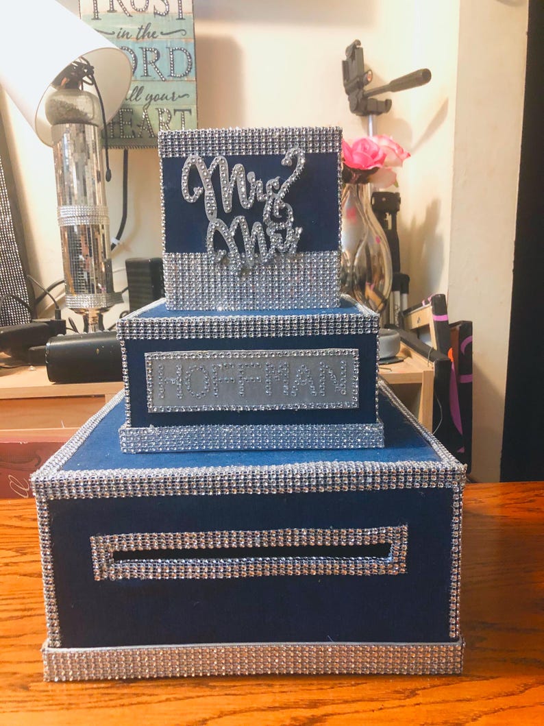 Puede incluir: Una caja de tarjetas de boda azul marino de tres niveles con ribete de diamantes de imitaci&oacute;n plateados. El nivel superior tiene "Mrs & Mrs" en escritura plateada. El nivel medio tiene el nombre "HOFFMAN" en plata.
