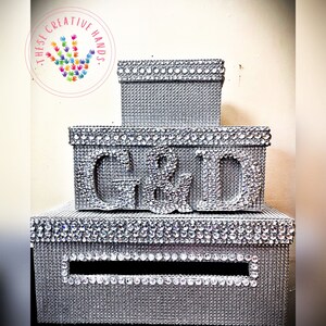 Puede incluir: Una pila de tres cajas plateadas, cada una adornada con brillantes pedrer&iacute;a. La caja del medio presenta las letras "C & D" en pedrer&iacute;a. La caja inferior tiene una ranura. Las cajas son probablemente para un evento especial.