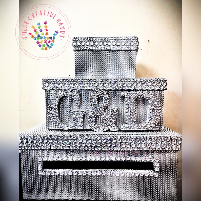 Puede incluir: Una pila de tres cajas plateadas, cada una adornada con brillantes pedrer&iacute;a. La caja del medio presenta las letras "C & D" en pedrer&iacute;a. La caja inferior tiene una ranura. Las cajas son probablemente para un evento especial.