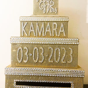 Puede incluir: Una caja de tarjetas de boda dorada de varios niveles con detalles de pedrer&iacute;a. La caja est&aacute; decorada con los nombres "Kamara", la fecha "03-03-2023" y las palabras "Mr & Mrs" en pedrer&iacute;a.