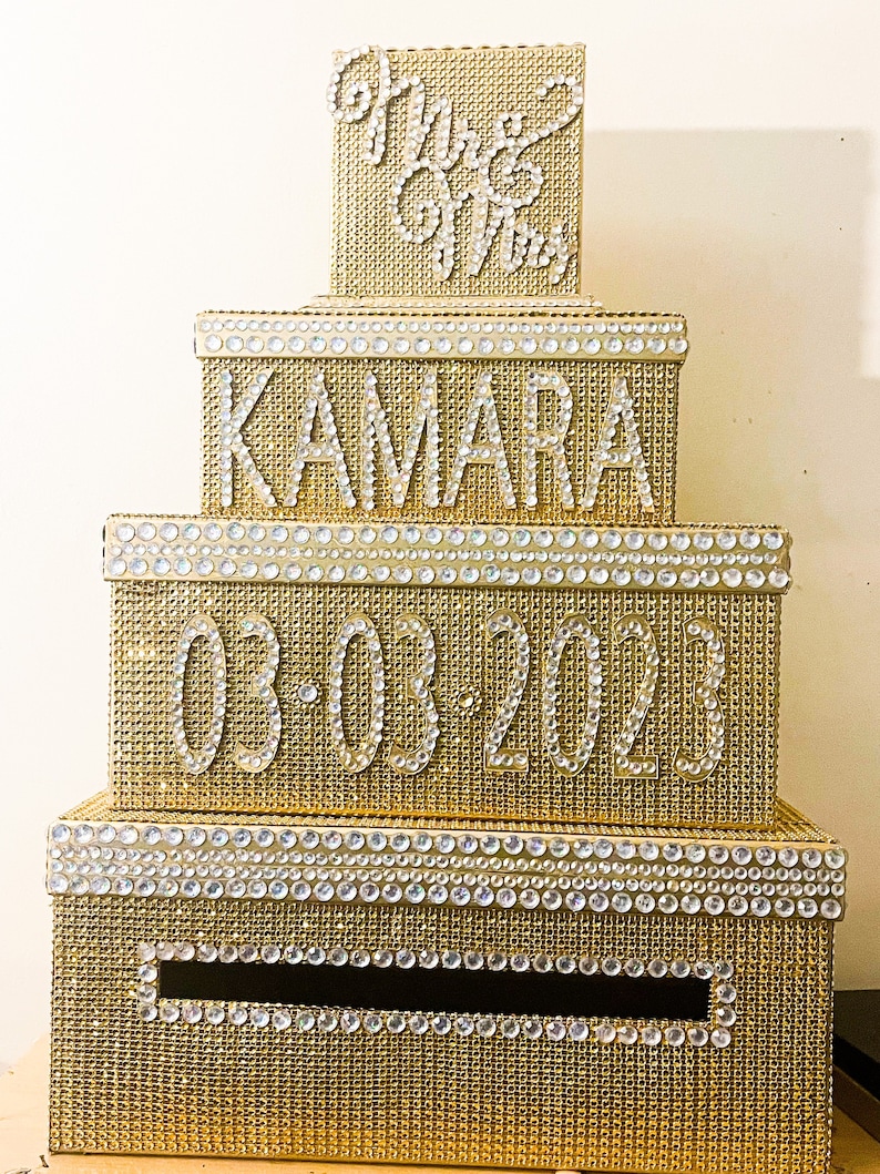 Puede incluir: Una caja de tarjetas de boda dorada de varios niveles con detalles de pedrer&iacute;a. La caja est&aacute; decorada con los nombres "Kamara", la fecha "03-03-2023" y las palabras "Mr & Mrs" en pedrer&iacute;a.
