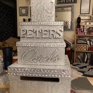 Puede incluir: Una caja de tarjetas de boda plateada de varios niveles. La caja est&aacute; cubierta de peque&ntilde;os pedrer&iacute;a brillantes. La parte superior dice "Mrs & Mrs", la del medio "Peters" y la tercera "Cards".