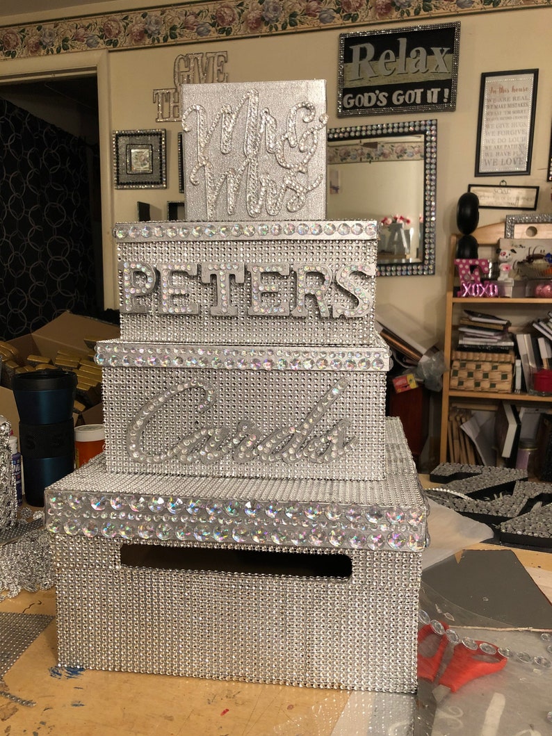 Puede incluir: Una caja de tarjetas de boda plateada de varios niveles. La caja est&aacute; cubierta de peque&ntilde;os pedrer&iacute;a brillantes. La parte superior dice "Mrs & Mrs", la del medio "Peters" y la tercera "Cards".