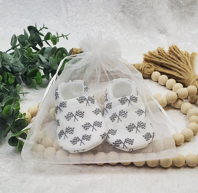 May include: A pair of white baby booties with a checkered flag pattern. The booties are in a white organza bag.