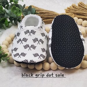May include: White baby booties with a black and white checkered flag pattern. The booties have a black grip dot sole.