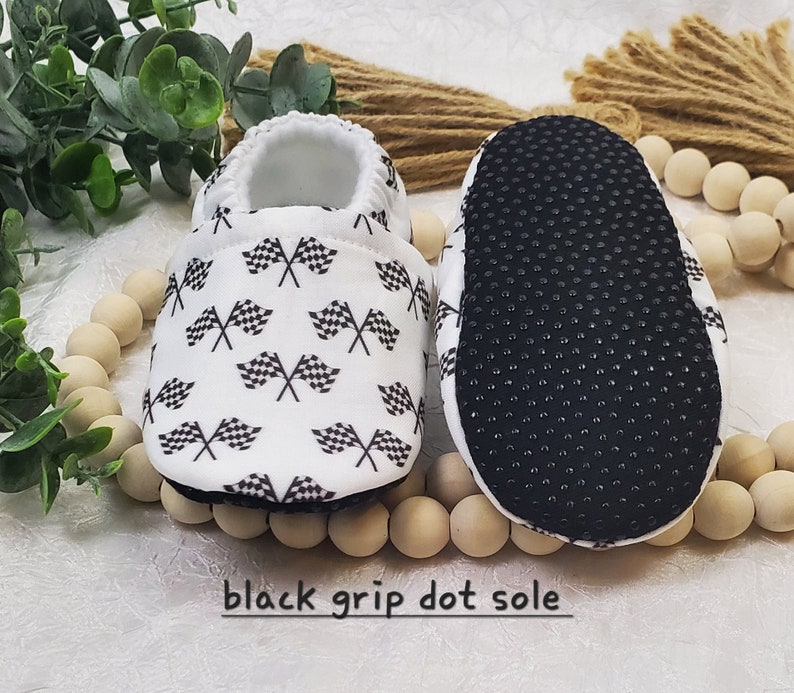 May include: White baby booties with a black and white checkered flag pattern. The booties have a black grip dot sole.