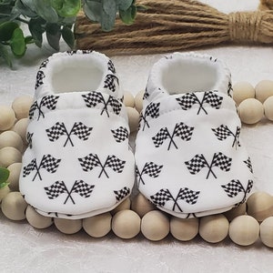 May include: A pair of white baby booties with a black and white checkered flag pattern.