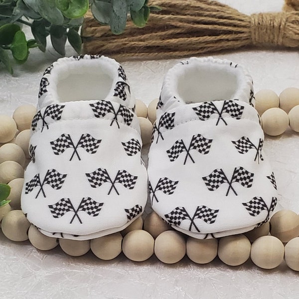 May include: A pair of white baby booties with a black and white chequered flag pattern.