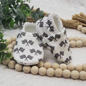 May include: A pair of white baby booties with a black and white checkered flag pattern. The booties are made of soft fabric and have a comfortable fit.