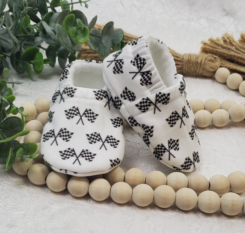 May include: A pair of white baby booties with a black and white checkered flag pattern. The booties are made of soft fabric and have a comfortable fit.