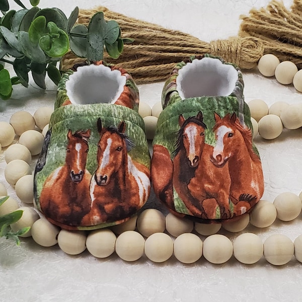 May include: A pair of baby shoes with a horse print. The shoes have a green and brown background with images of brown and white horses. The interior of the shoes is white. The shoes are made of soft fabric.