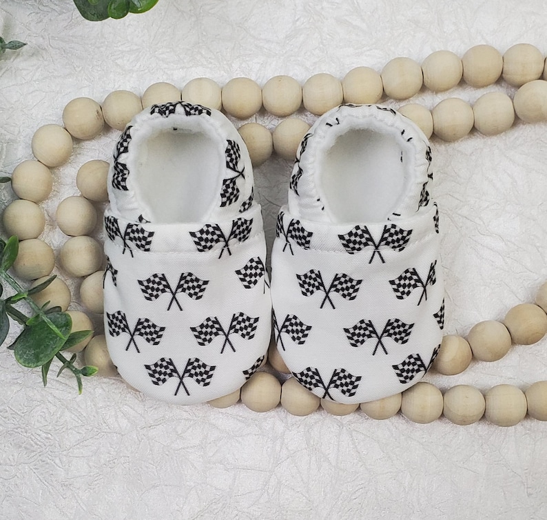 May include: A pair of white baby booties with a black and white checkered flag pattern.
