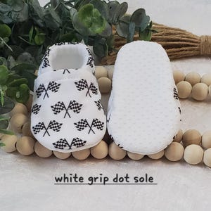 May include: White baby shoes with a checkered flag pattern and white grip dot soles. "white grip dot sole" is written on the bottom of the image.