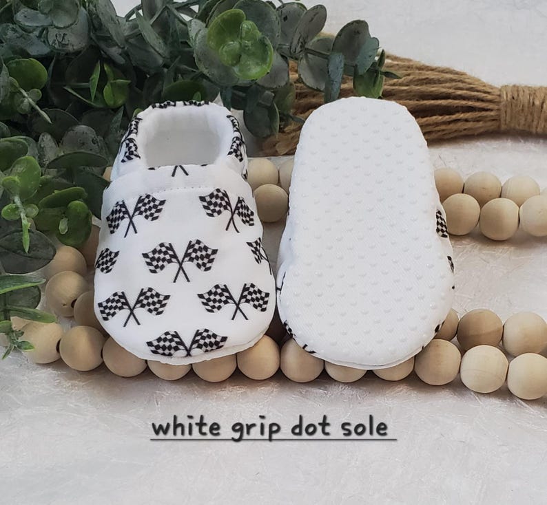 May include: White baby shoes with a checkered flag pattern and white grip dot soles. "white grip dot sole" is written on the bottom of the image.