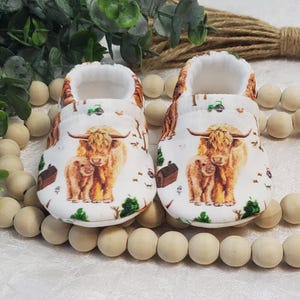 May include: A pair of white baby booties with a highland cow and calf print. The booties have a white background with brown, black, and white cows and calves. The booties are made of soft fabric and are perfect for keeping baby's feet warm and cozy.