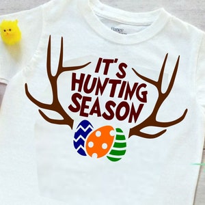 May include: White t-shirt with the text "IT'S HUNTING SEASON" in brown, with brown antlers and colorful Easter eggs. A basket of colorful eggs and toy chicks are also in the image.
