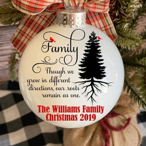 Op de afbeelding: Witte kerstbal met het woord "Family" in script, een zwarte boomafbeelding en de tekst "Though we grow in different directions, our roots remain as one." Een rood-witte geruite strik is bevestigd.